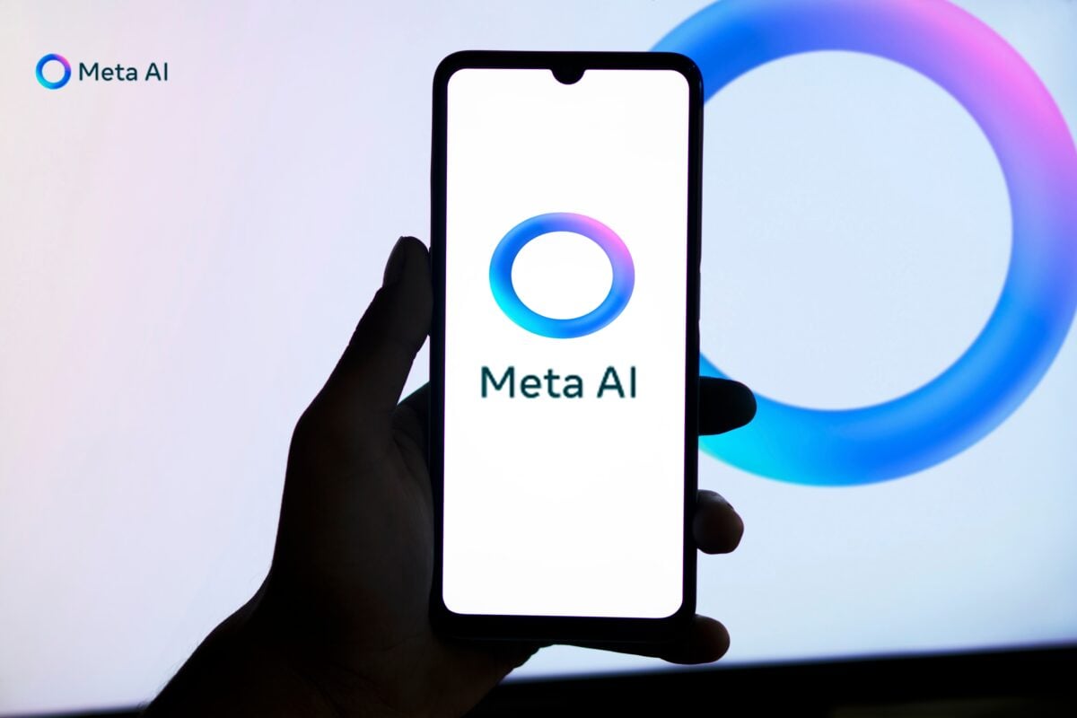 A hand holds a smartphone displaying the "Meta AI" logo against a blurred monitor featuring a similar circular design. Related: Meta Connect